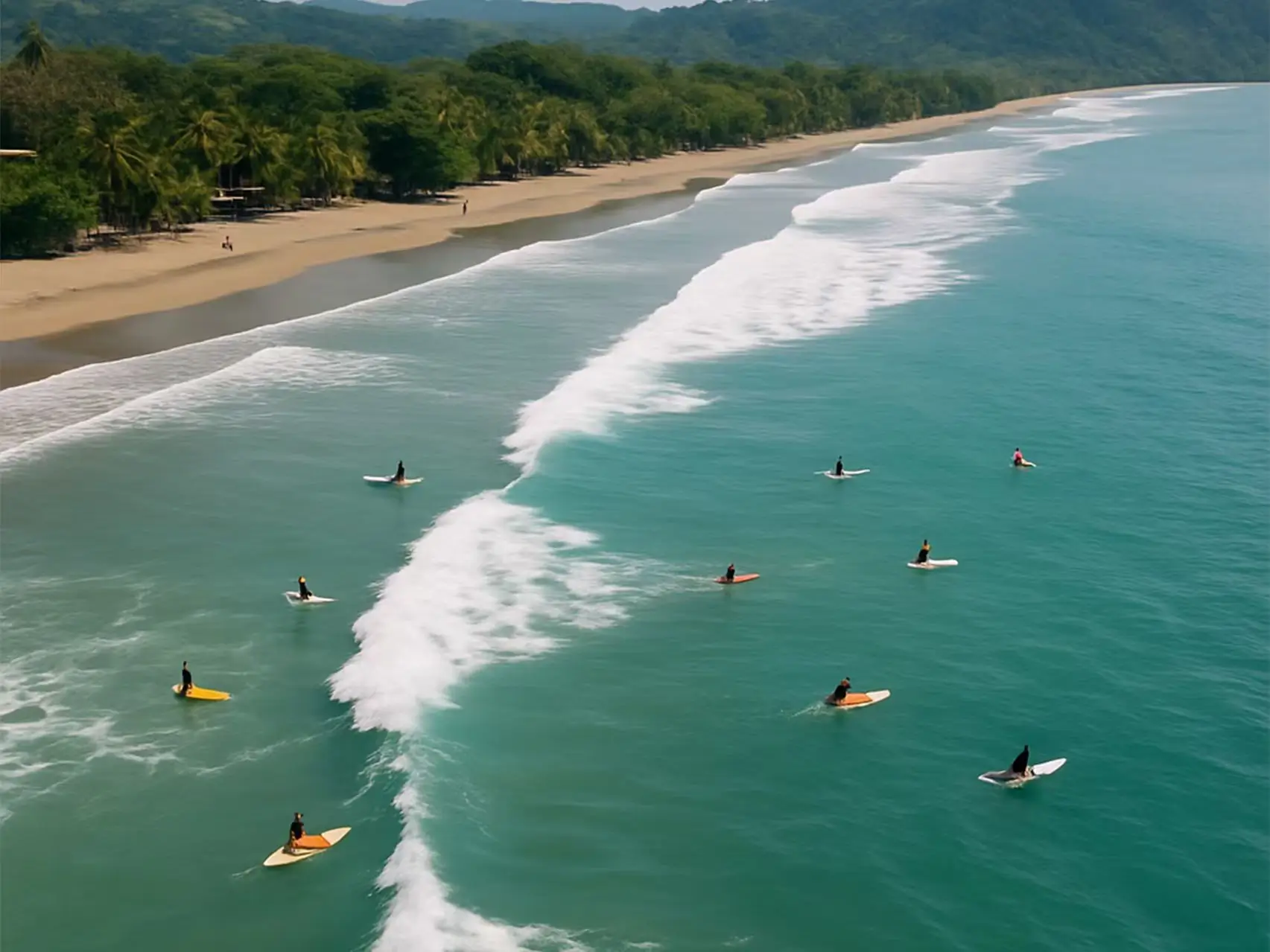 Aerial view of surfers waiting for waves in the tropical blue waters of Costa Rica, surrounded by a lush green rainforest coastline.
