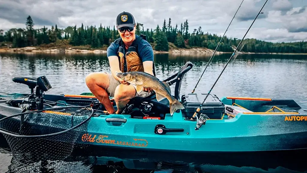 A person sitting in a turquoise fishing kayak, holding a large fish, with fishing rods and a trolling motor visible, demonstrating hands-free fishing capabilities on a lake.