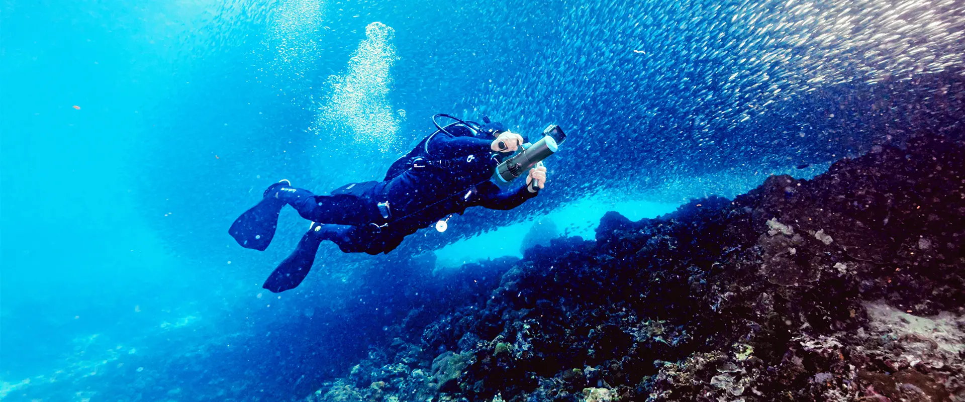 Wide desktop banner showing a diver using the handheld EDGE K5 underwater scooter to glide alongside a massive school of fish over a deep coral reef. Perfect for both scuba diving and snorkeling adventures.
